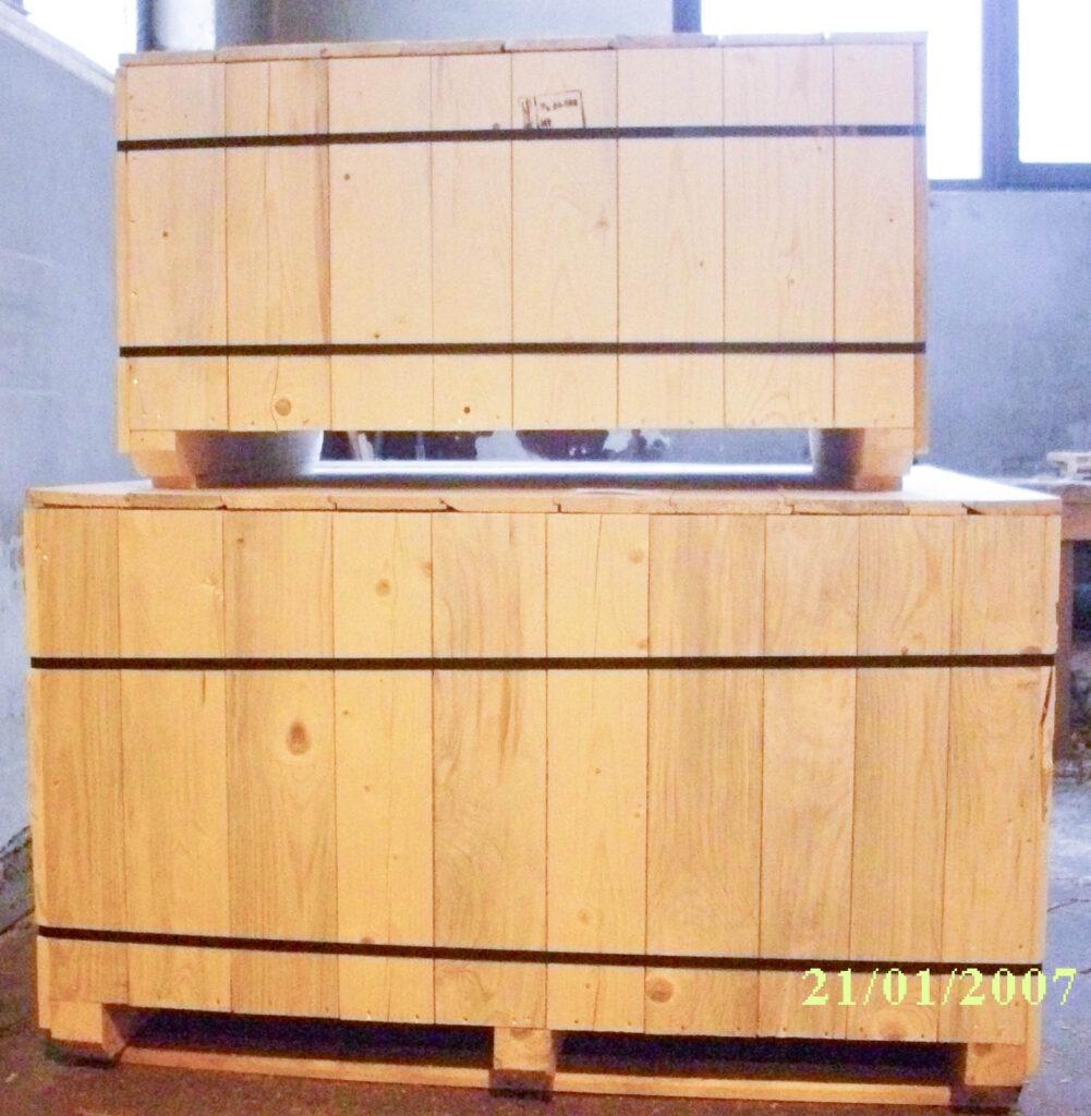 Dwie drewniane skrzynie transportowe, jedna na drugiej, zabezpieczone czarnymi pasami, widoczny fragment podłogi i okno w tle.