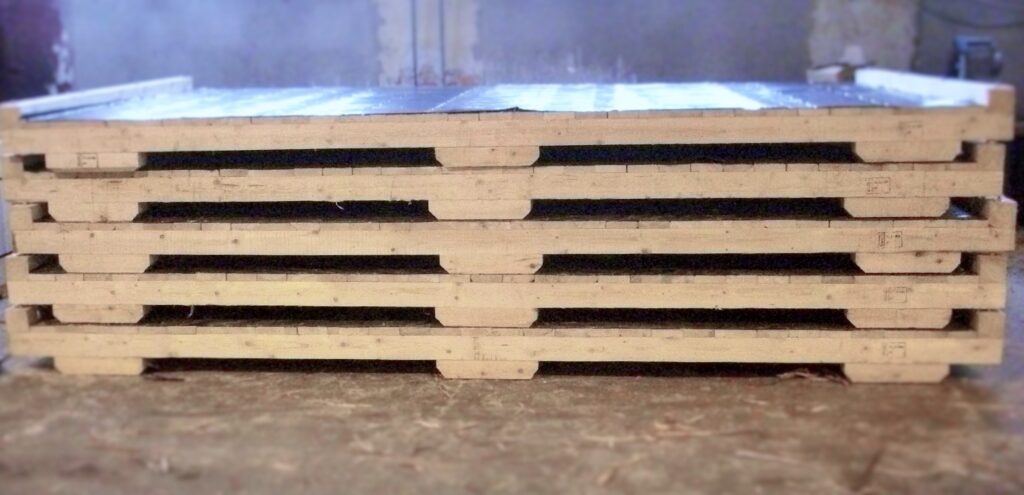 Stos drewnianych palet z widocznymi wgłębieniami w deskach, zabezpieczonych folią na górze, leżących na betonowej podłodze w hali.