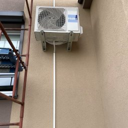 FK Klimatyzacje - Zewnętrzna jednostka klimatyzacji Haier zamontowana na ścianie budynku, widoczne rury i kable, obok rusztowanie.