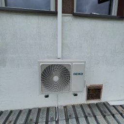 FK Klimatyzacje - Zewnętrzna jednostka klimatyzacji marki HEIKO zamontowana na jasnej, teksturowanej ścianie budynku, widoczne okna powyżej i metalowa osłona poniżej.