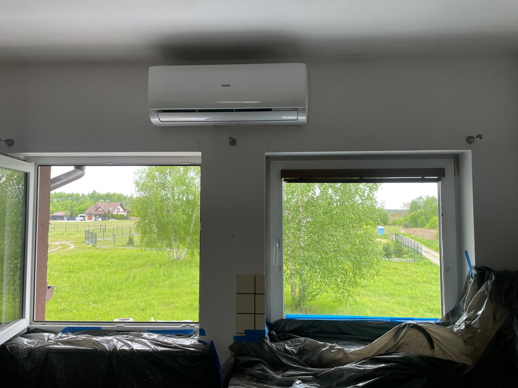 Zamontowany klimatyzator marki HEIKO na ścianie w pomieszczeniu z dwoma oknami zabezpieczonymi folią malarską, widok na zielony ogród i dom.