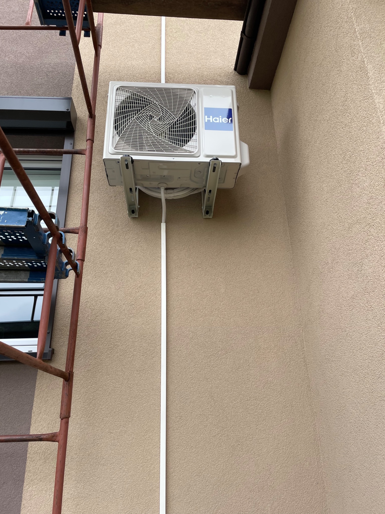 Zewnętrzna jednostka klimatyzacji Haier zamontowana na ścianie budynku, widoczne rury i kable, obok rusztowanie.