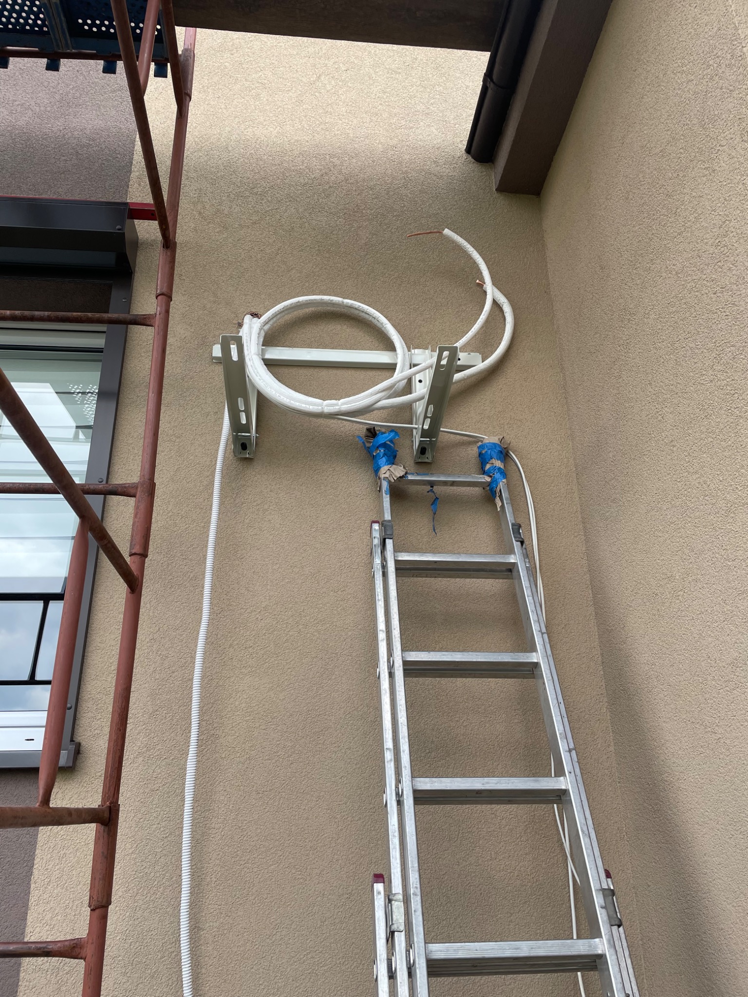 Montaż klimatyzacji: rury i przewody zamocowane na wspornikach na elewacji budynku, drabina przystawiona do ściany, widoczne rusztowanie.