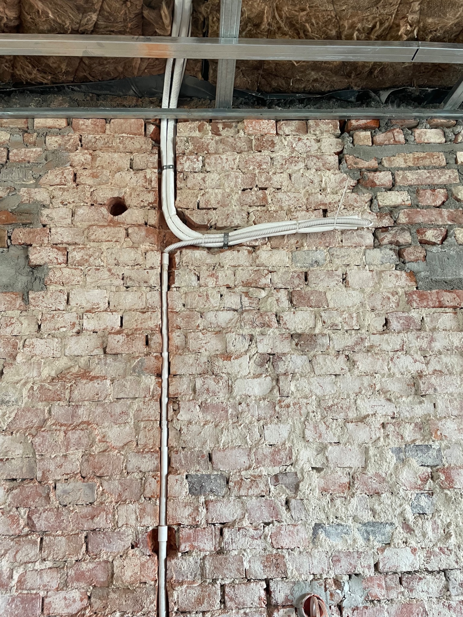 Odsłonięta ceglana ściana z poprowadzonymi rurami instalacyjnymi i fragmentem konstrukcji sufitu podwieszanego z izolacją.