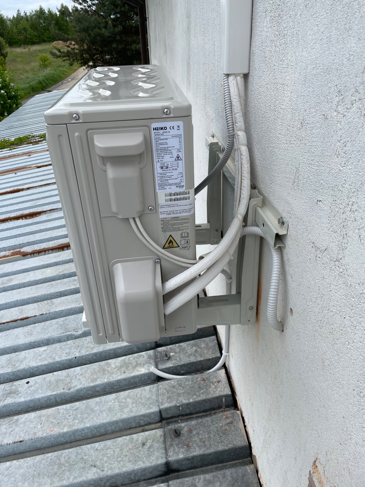 Zewnętrzna jednostka klimatyzacji marki Heiko zamontowana na metalowym dachu przy białej ścianie budynku, z widocznymi przewodami w izolacji.