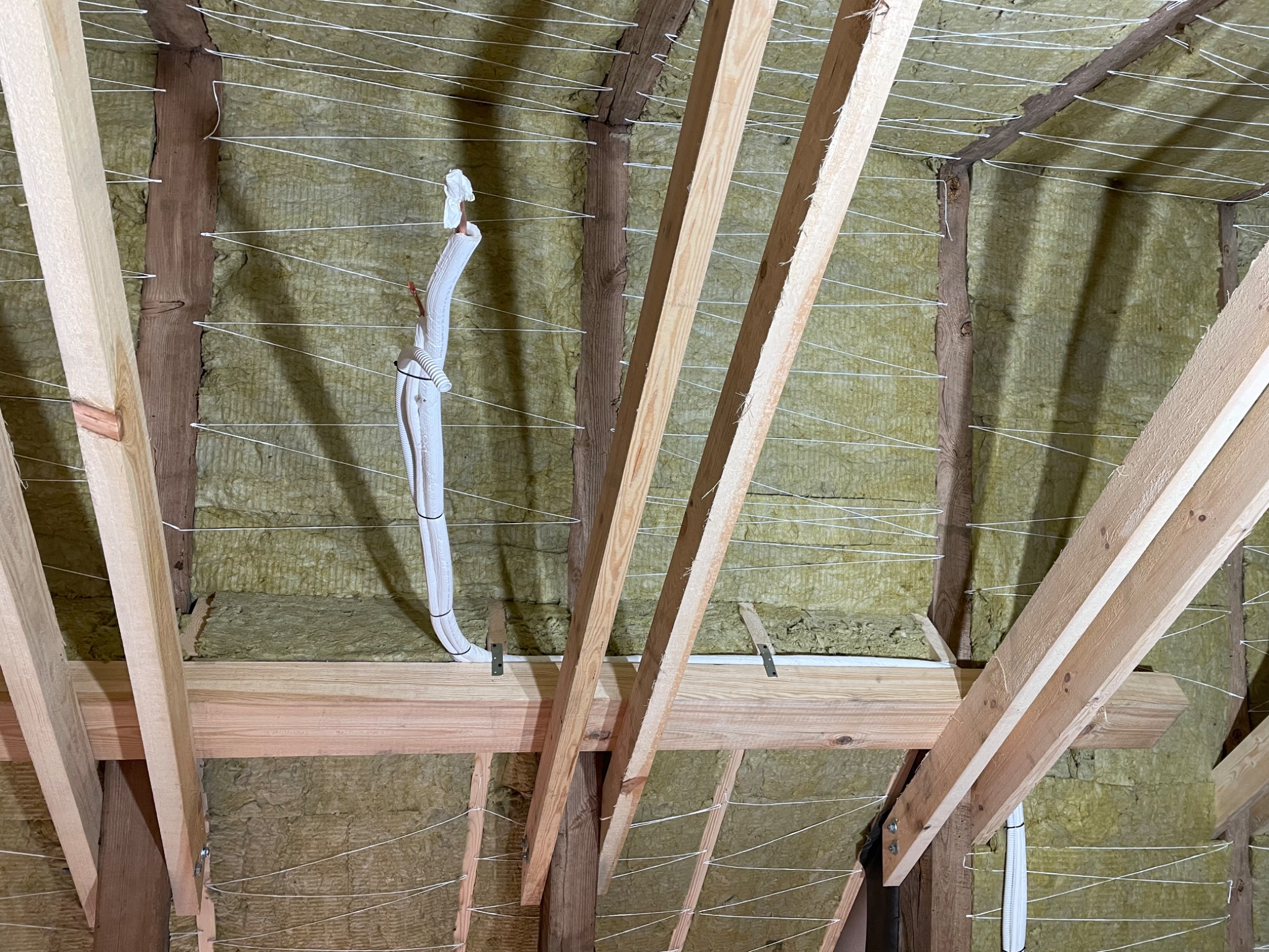 Montaż instalacji w poddaszu z widoczną izolacją, drewnianą konstrukcją dachu i rurami instalacji klimatyzacyjnej przymocowanymi sznurkiem.