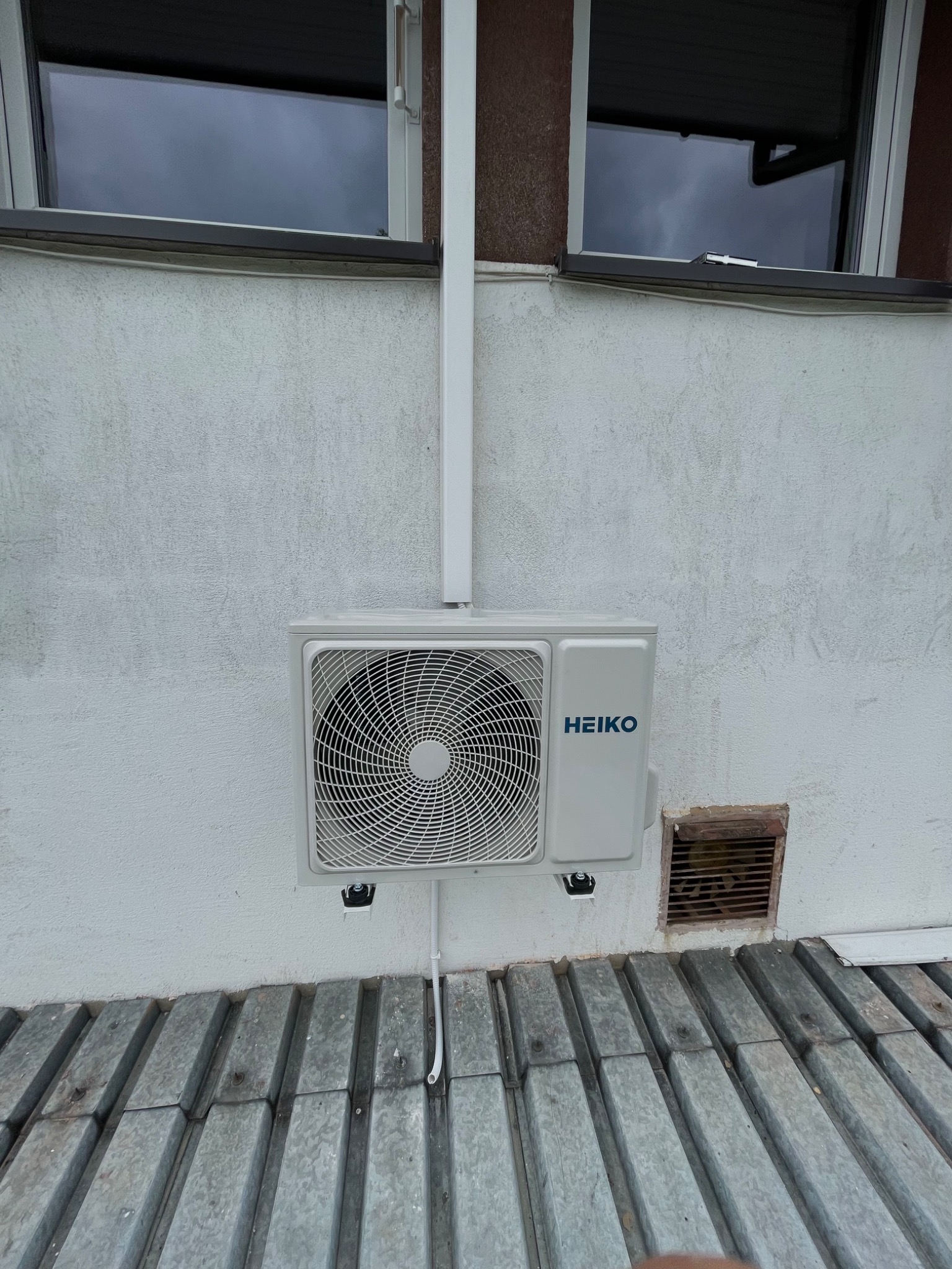 Zewnętrzna jednostka klimatyzacji marki HEIKO zamontowana na jasnej, teksturowanej ścianie budynku, widoczne okna powyżej i metalowa osłona poniżej.
