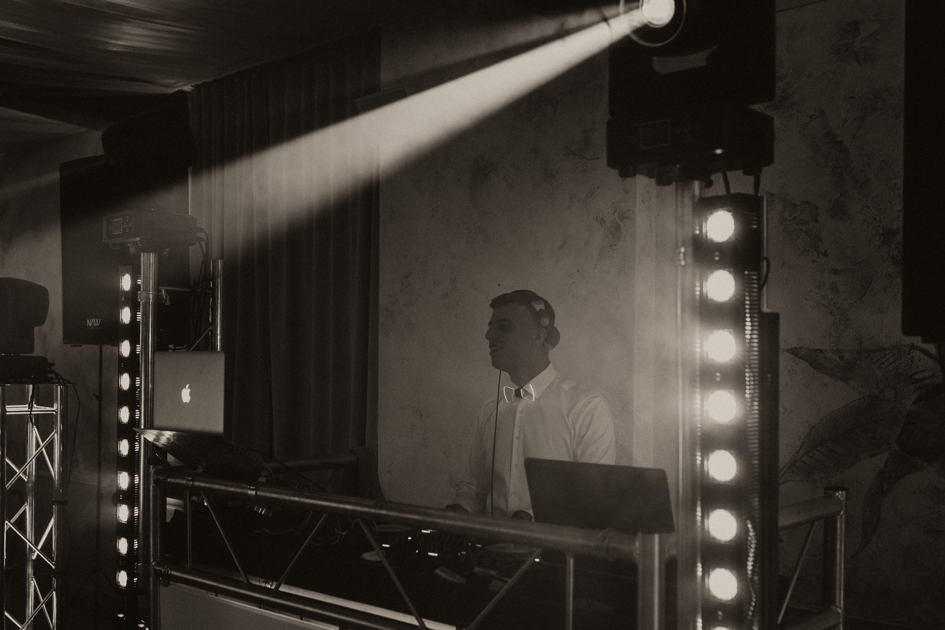 DJ w białej koszuli i podświetlaną muszką, za konsoletą z laptopami, oświetlony punktowym światłem reflektora, w tle kolumny głośnikowe i dekoracyjna ściana.