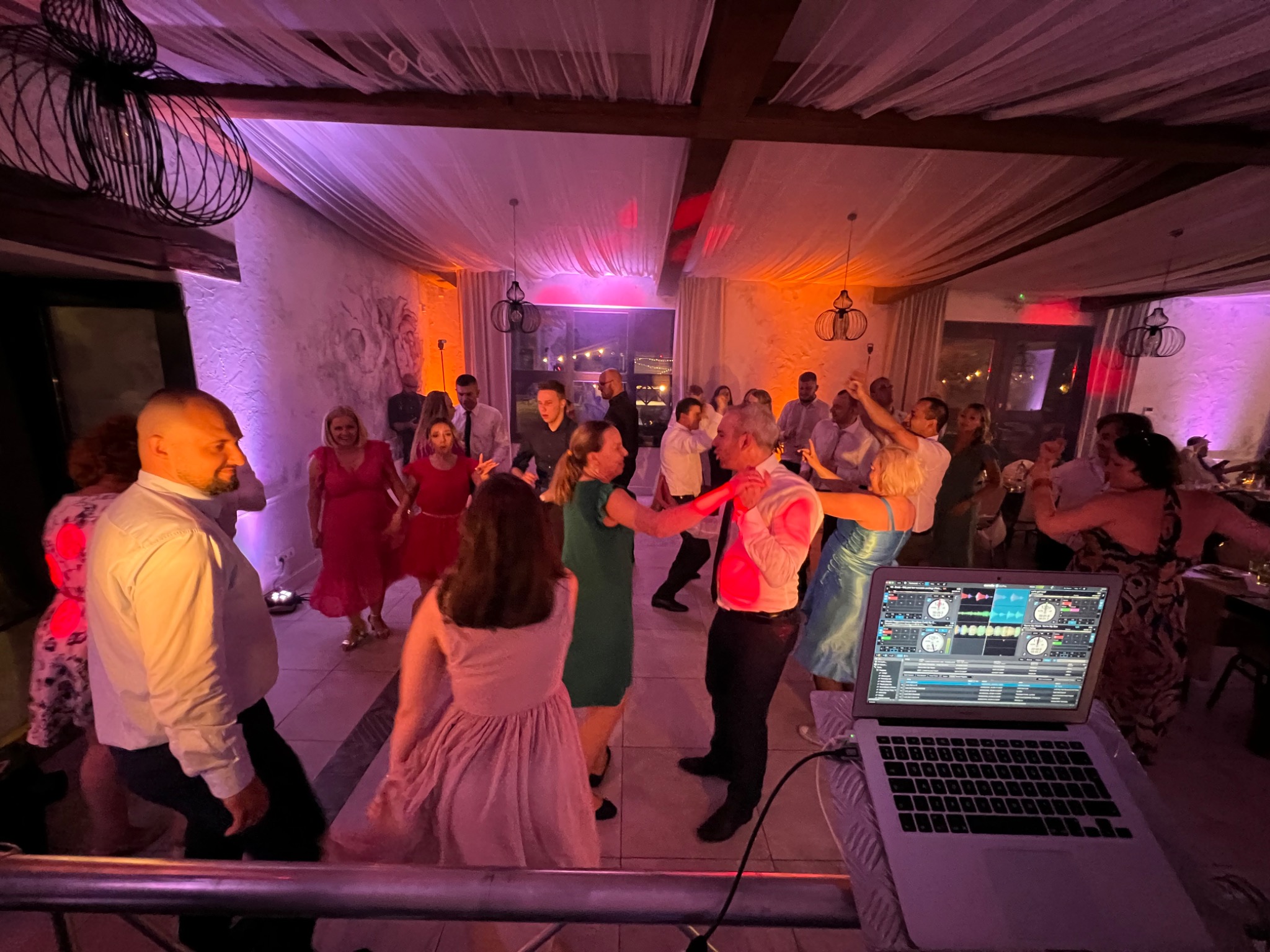 Tańczący goście weselni w sali z dekoracjami, oświetleni kolorowym światłem, z laptopem z oprogramowaniem DJ-skim na pierwszym planie.