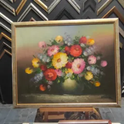 Olejny obraz z bukietem kolorowych kwiat&oacute;w w ozdobnej, złotej ramie, ustawiony na drewnianym stojaku, w tle ekspozycja r&oacute;żnorodnych ram do obraz&oacute;w.