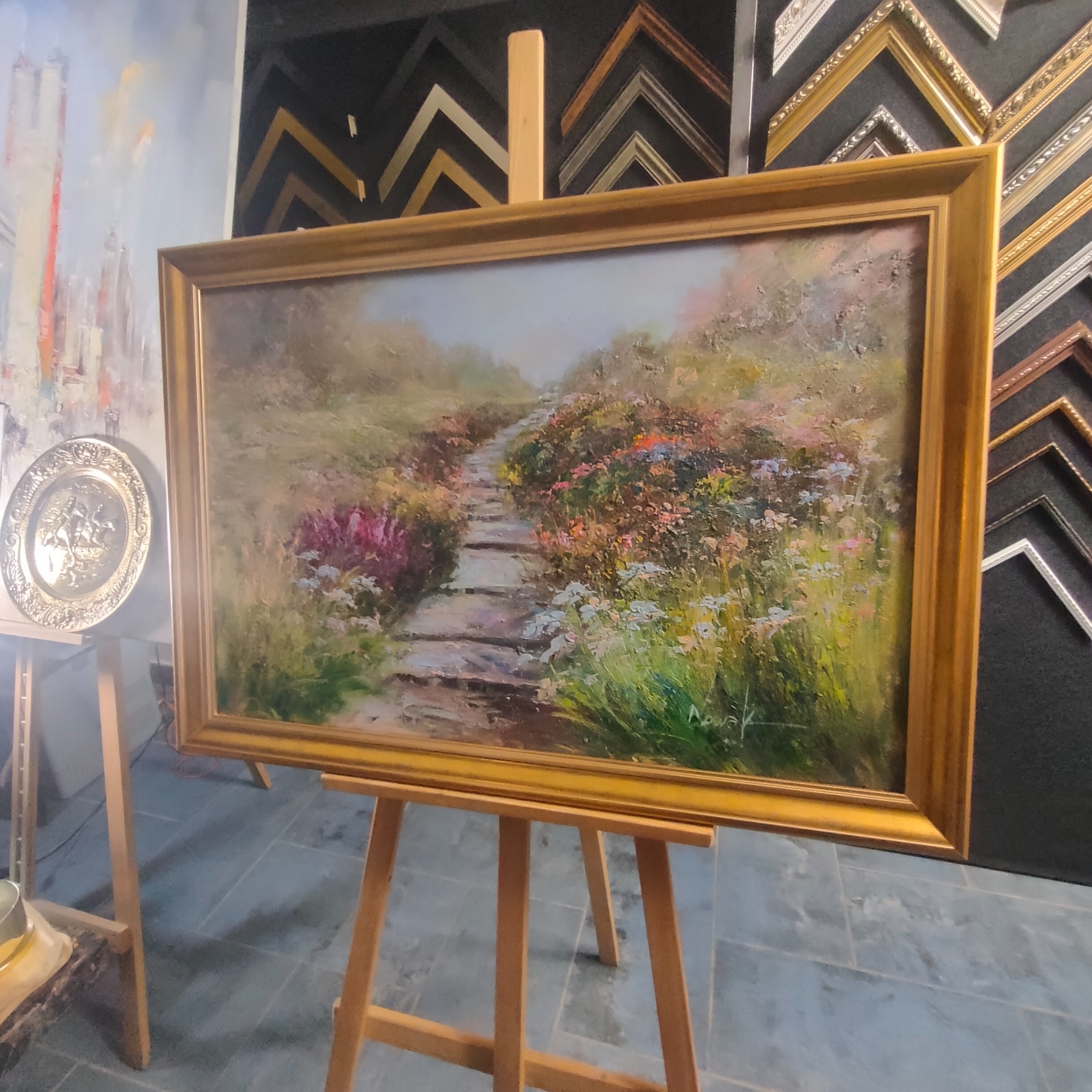 Obraz olejny przedstawiający ścieżkę w ogrodzie, oprawiony w złotą ramę i umieszczony na drewnianym sztaludze; w tle ściana z ekspozycją ramek do obrazów.