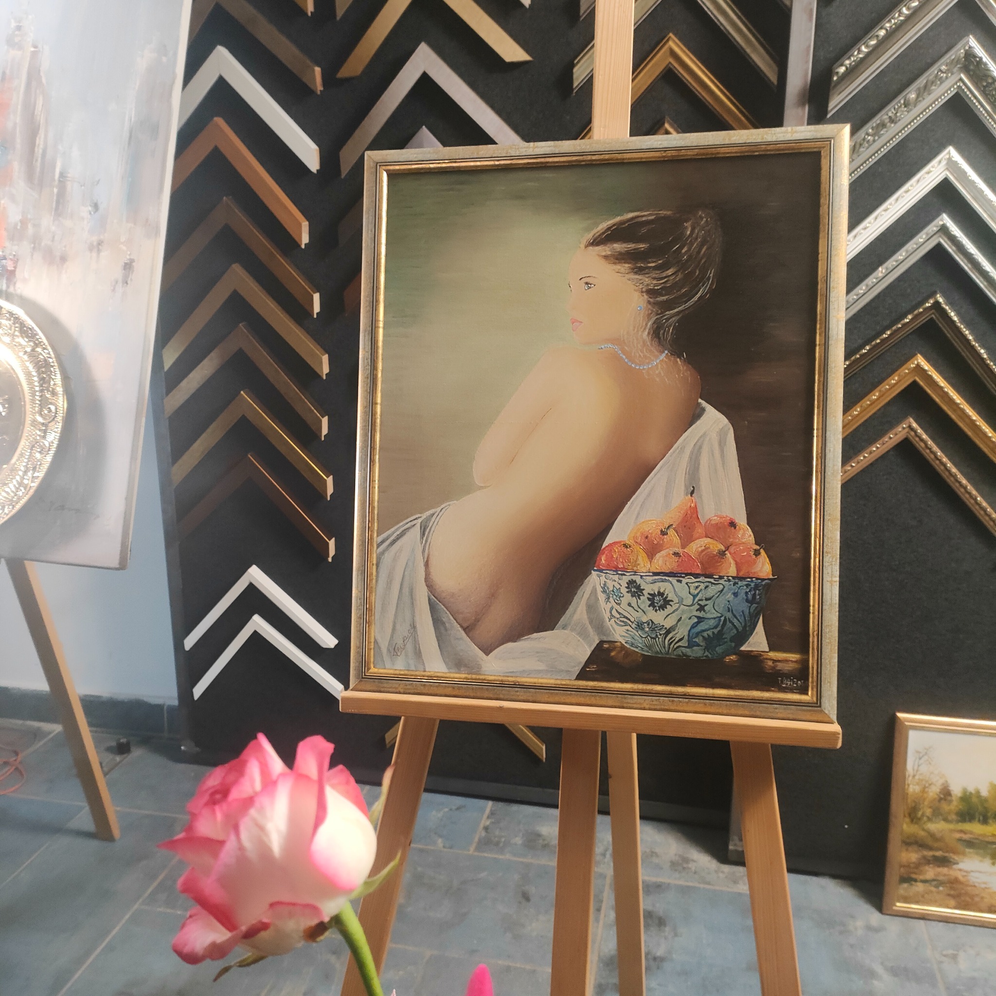 Obraz przedstawiający akt kobiety z misą owoców w złotej ramie, ustawiony na drewnianej sztaludze w pracowni z ekspozycją ram, z różową różą w wazonie na pierwszym planie.