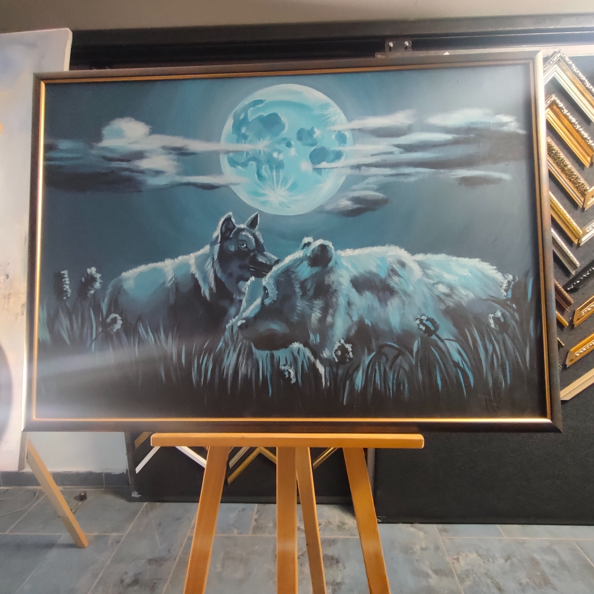 Oprawiony obraz przedstawiający niedźwiedzia i wilka na tle księżyca, umieszczony na drewnianym stojaku w pracowni z ramami w tle.
