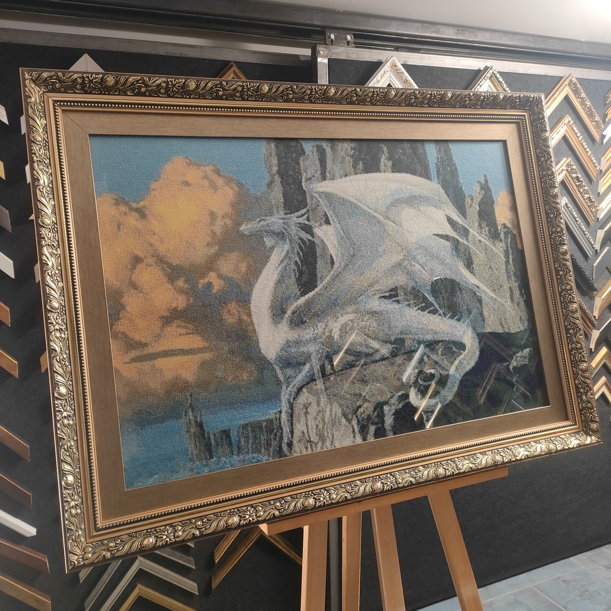 Oprawiony w złotą, zdobioną ramę haft krzyżykowy przedstawiający białego smoka na tle skalistego krajobrazu i zachodzącego słońca, ustawiony na drewnianym stojaku w pracowni szklarza.