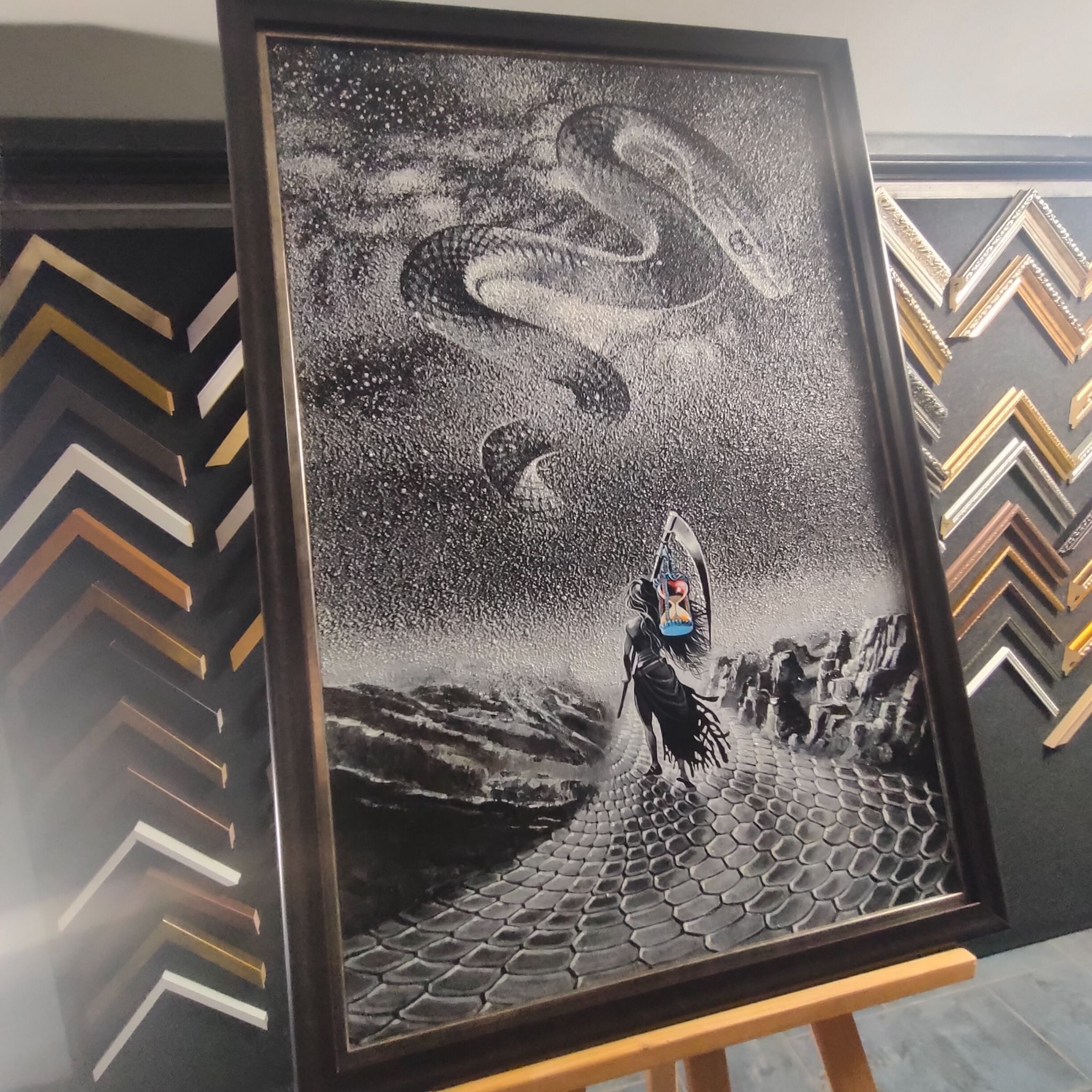 Oprawiony w ciemną ramę obraz przedstawiający postać z kosą i klepsydrą na tle krajobrazu z wężem na niebie, widoczne próbki ram na ścianie.