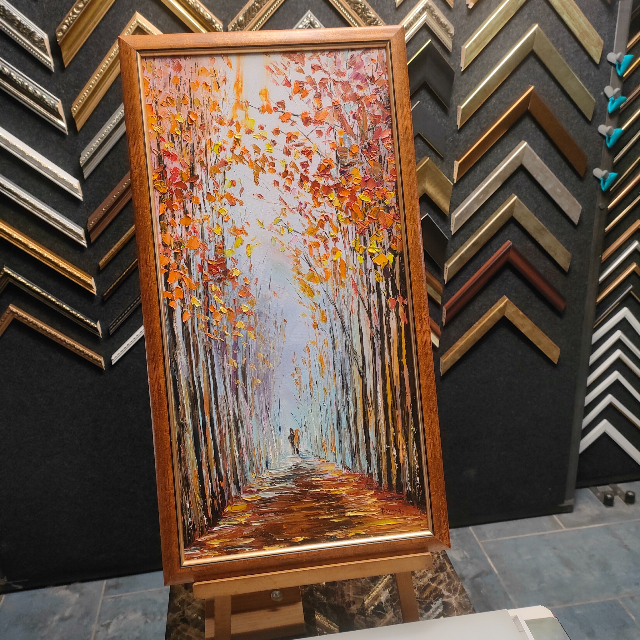 Obraz olejny w drewnianej ramie przedstawiający jesienny pejzaż z parą spacerującą pośród drzew, eksponowany na sztaludze w pracowni z różnymi ramami w tle.