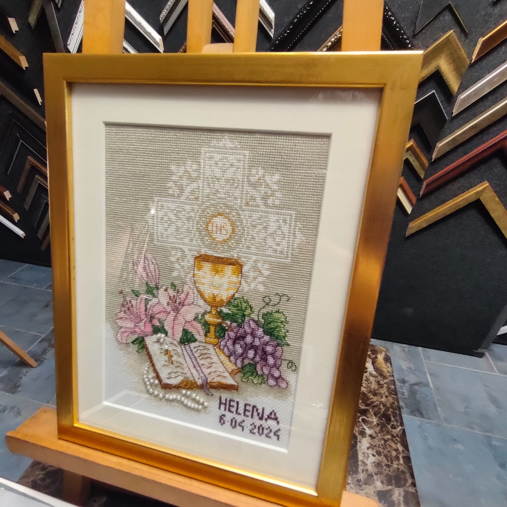 Haft krzyżykowy z motywem religijnym, oprawiony w złotą ramę, stojący na drewnianym stojaku; w tle widoczne próbki ram.