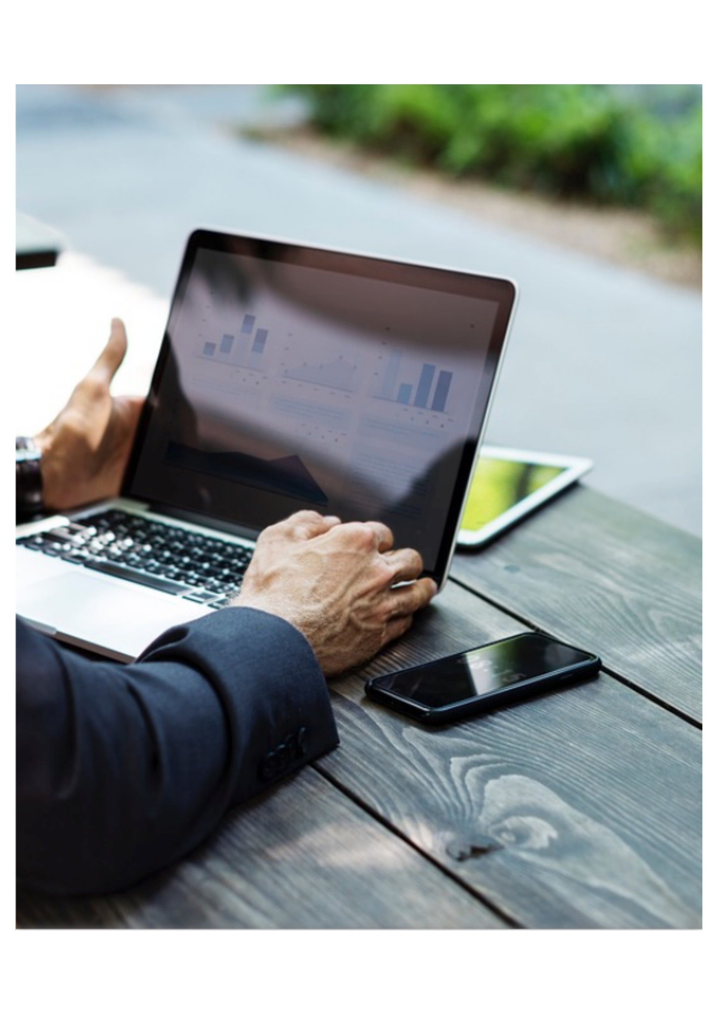 Mężczyzna w garniturze analizuje wykresy na laptopie na drewnianym stole, obok leży tablet i smartfon.