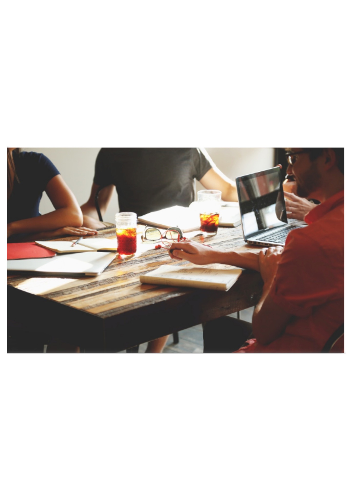 Grupa osób przy drewnianym stole, na którym znajdują się notatniki, laptop, okulary i napoje w słoikach, podczas spotkania w jasnym pomieszczeniu.