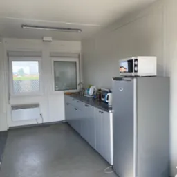 Wnętrze kontenera biurowego z aneksem kuchennym: szare szafki, lodówka z mikrofalówką, okna z widokiem na zewnątrz.