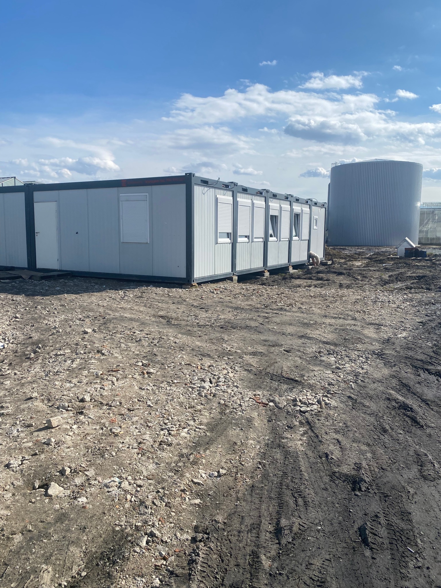 Zestaw połączonych, szarych kontenerów biurowych z roletami, ustawionych na placu budowy z widocznym zbiornikiem przemysłowym w tle.