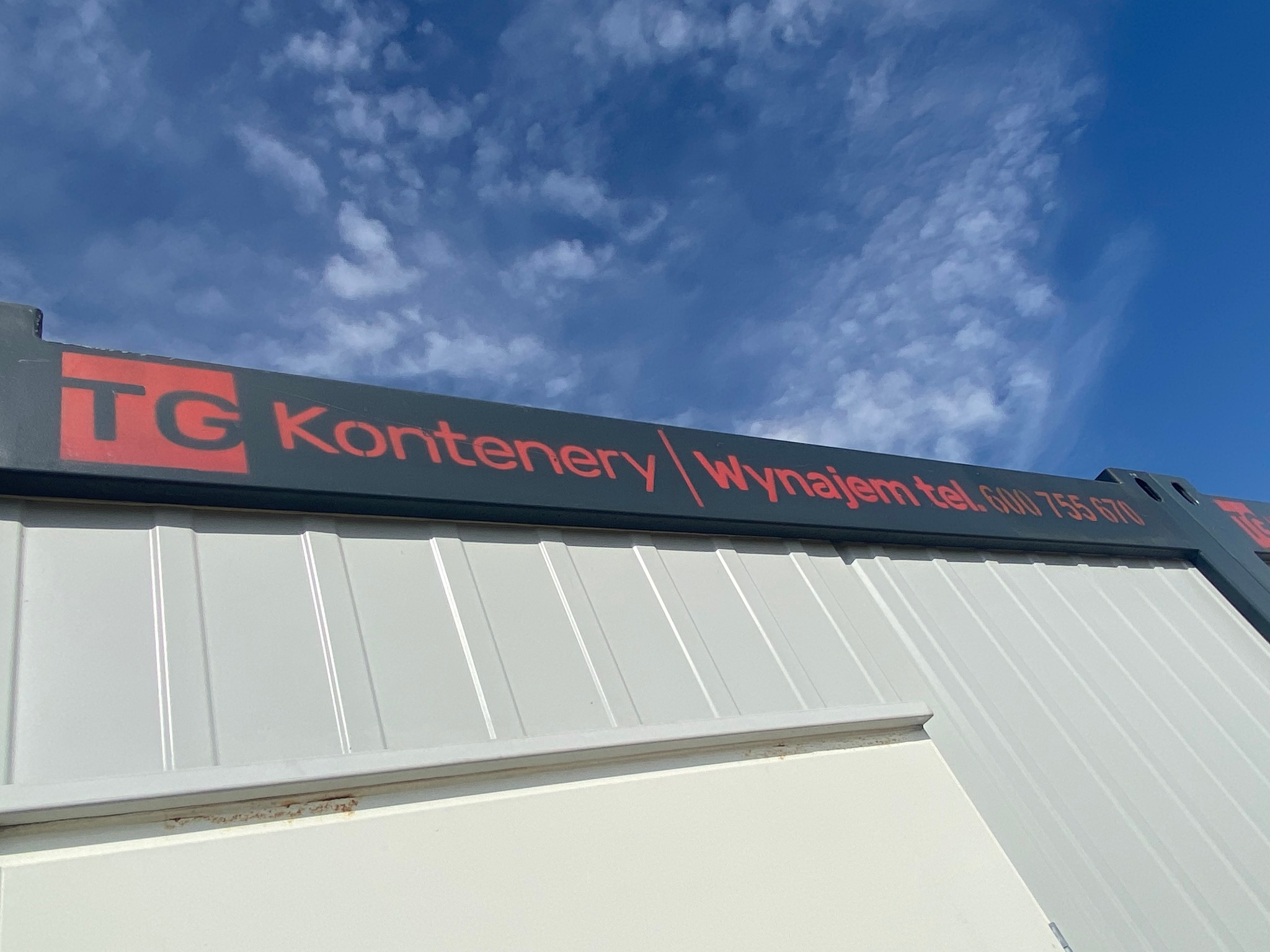 Biały kontener z czerwonym napisem 'TG Kontenery / Wynajem tel. 600 755 670' na tle błękitnego nieba z chmurami.