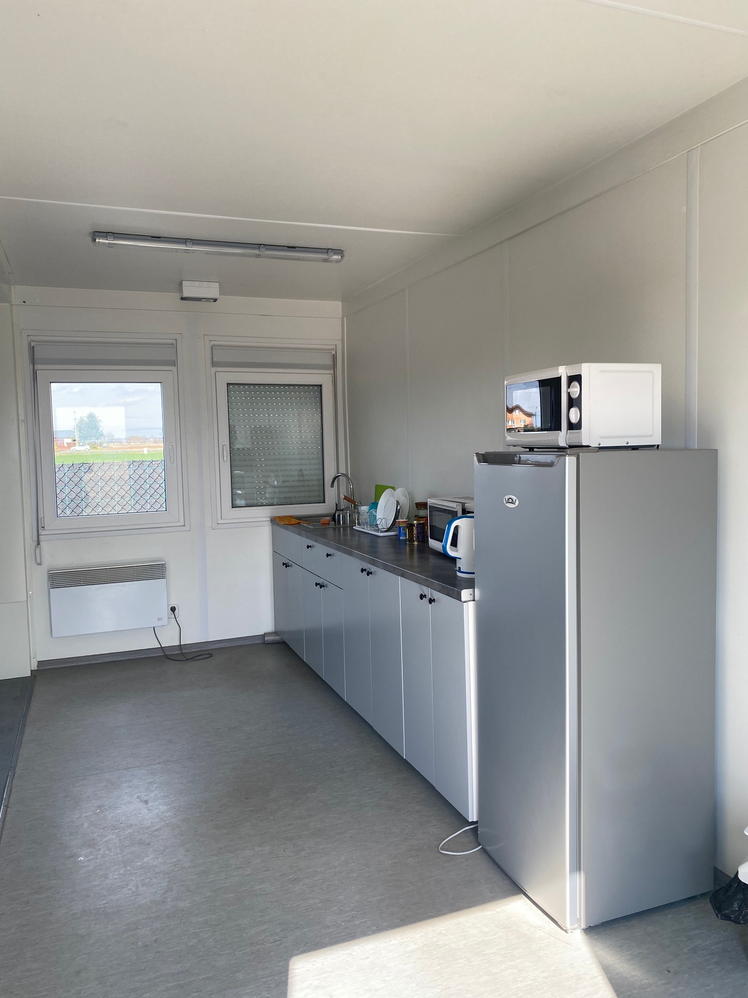 Wnętrze kontenera biurowego z aneksem kuchennym: szare szafki, lodówka z mikrofalówką, okna z widokiem na zewnątrz.