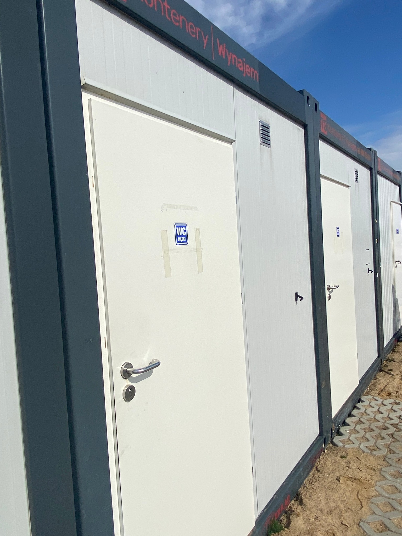 Rząd przenośnych toalet w kontenerach z białymi drzwiami i niebieskimi znakami WC, ustawionych na placu budowy pod błękitnym niebem.