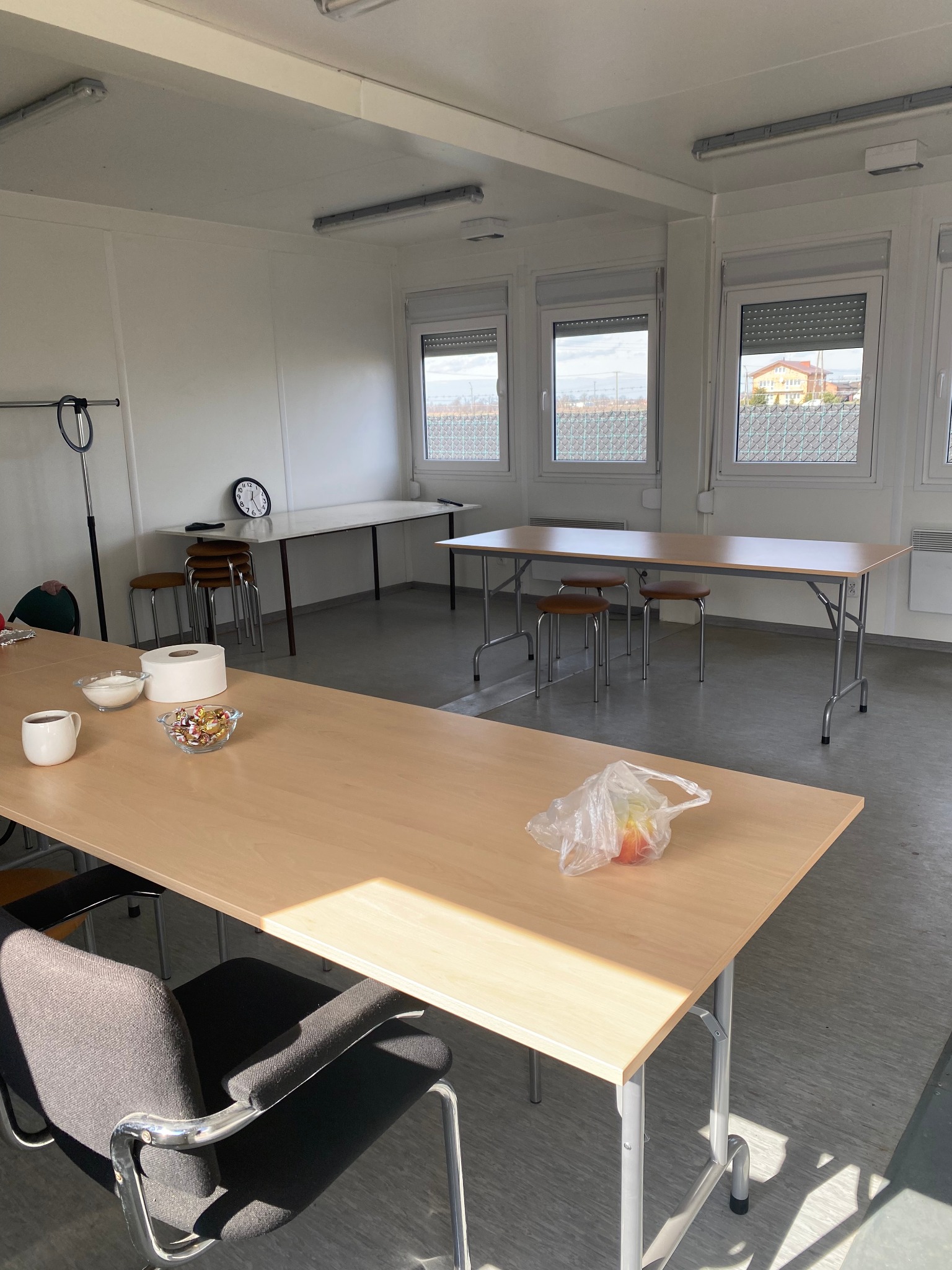 Wnętrze kontenera biurowego z trzema oknami, stołami i krzesłami. Na stole leżą słodycze, cukier, ręcznik papierowy i jabłka w foliowej torebce.