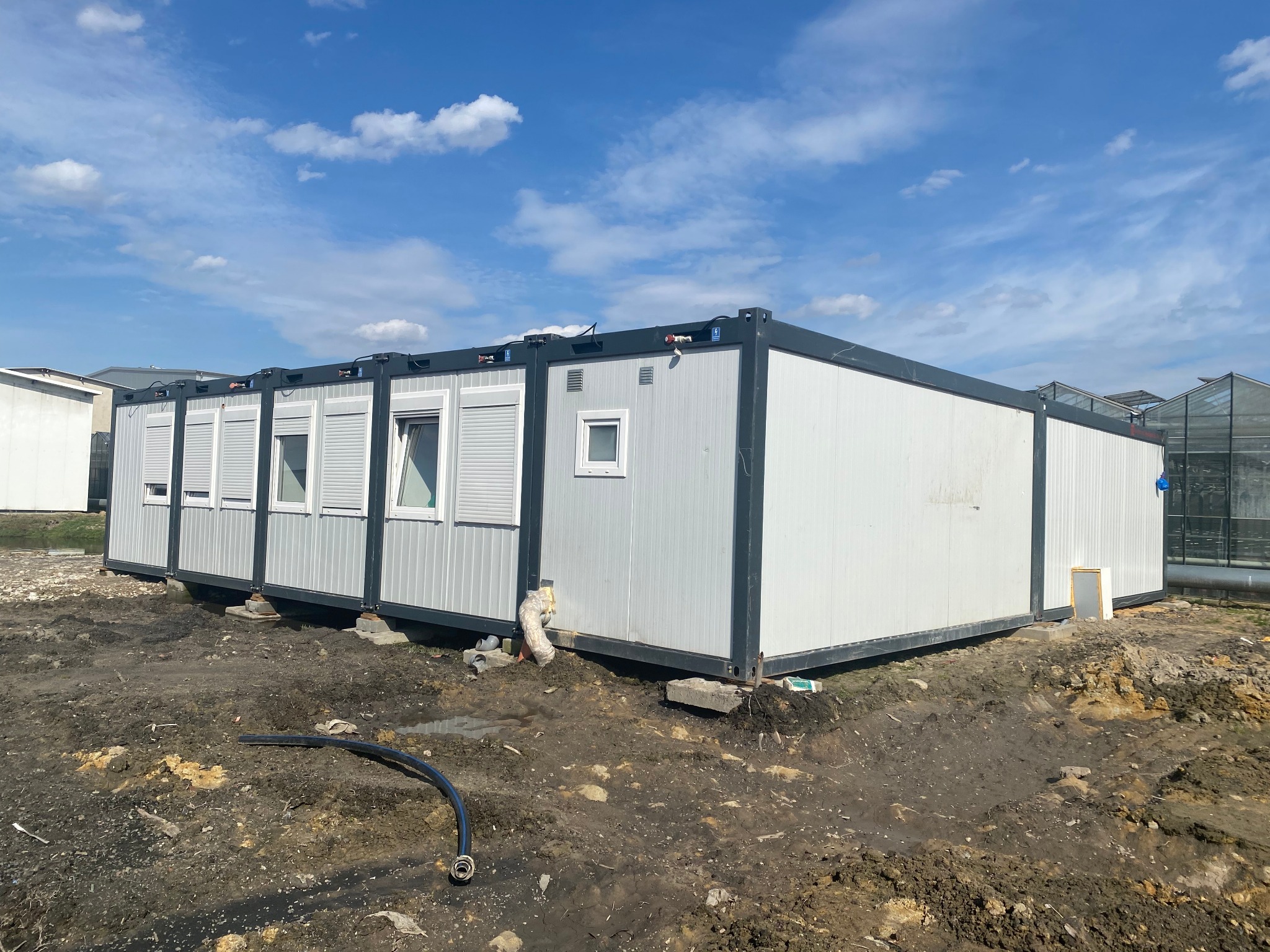 Zestaw połączonych kontenerów biurowych z oknami i roletami na placu budowy, widoczny wąż doprowadzający wodę i uprawa szklarniowa w tle.