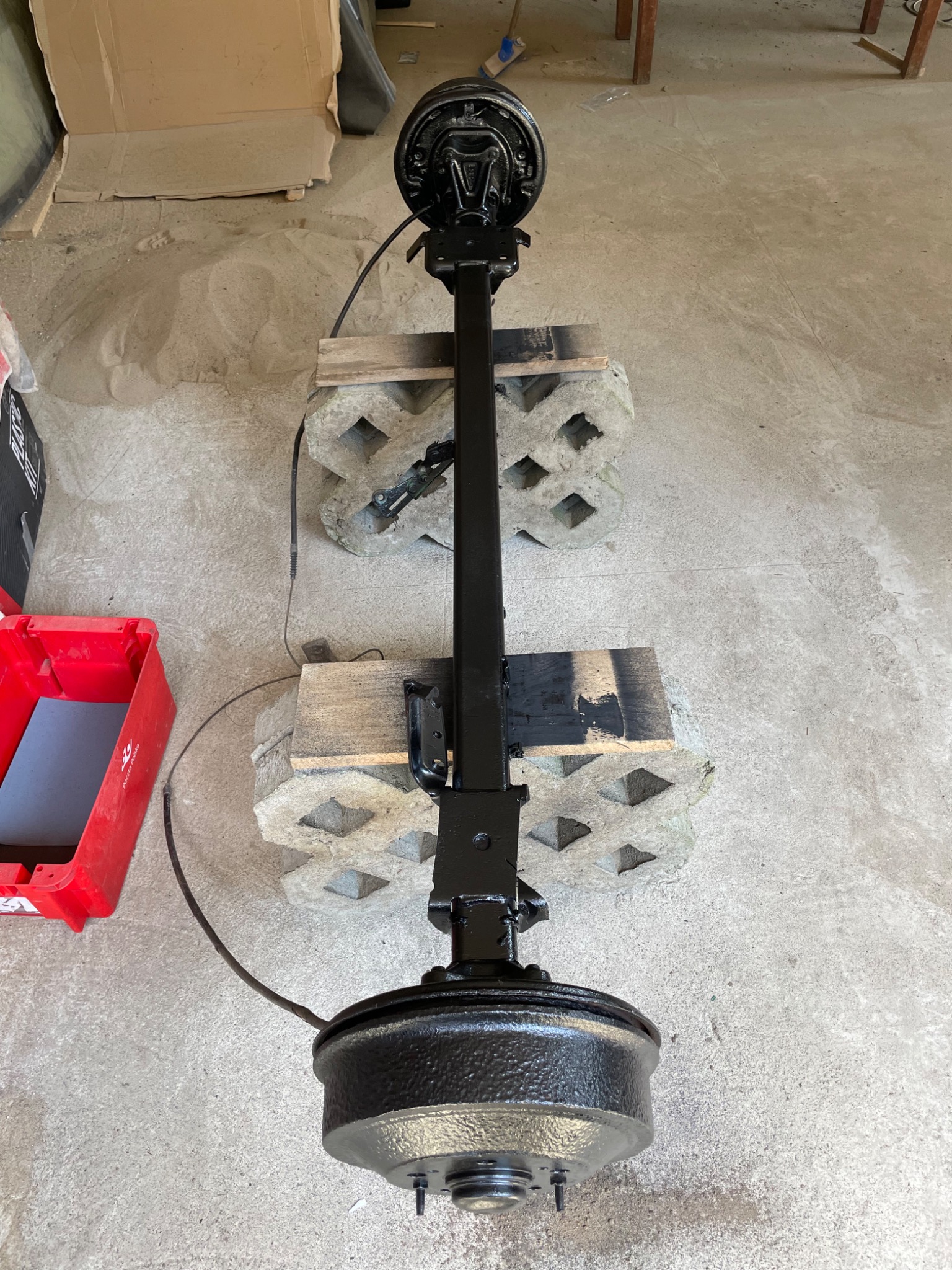 Tylna oś pojazdu po renowacji, pomalowana na czarno, wsparta na betonowych bloczkach. Widoczny przewód hamulcowy i ślady piasku.