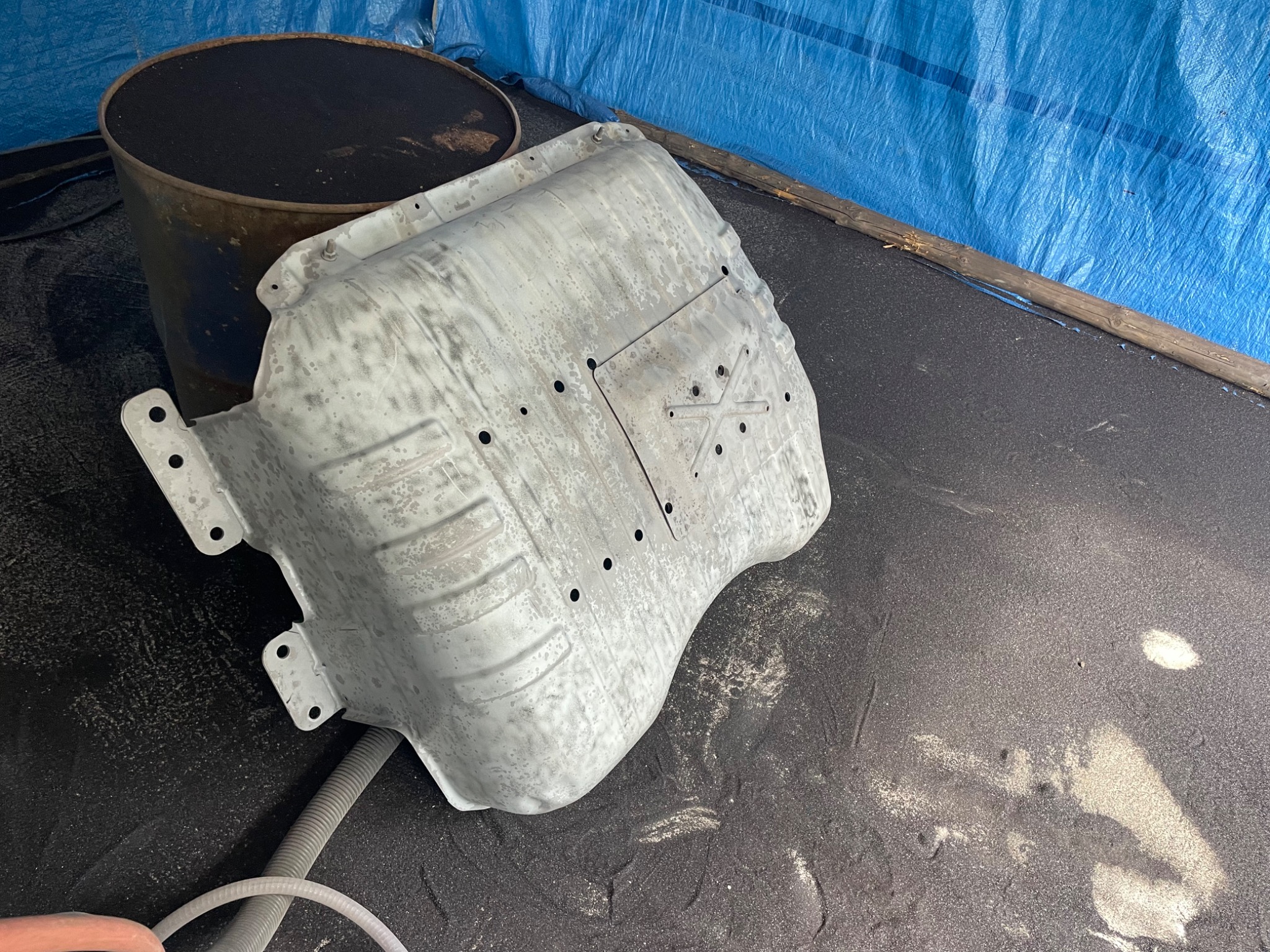 Metalowa osłona po obróbce ściernej, widoczne ślady wcześniejszej korozji i resztki materiału ściernego na powierzchni, obok stoi beczka i wąż.