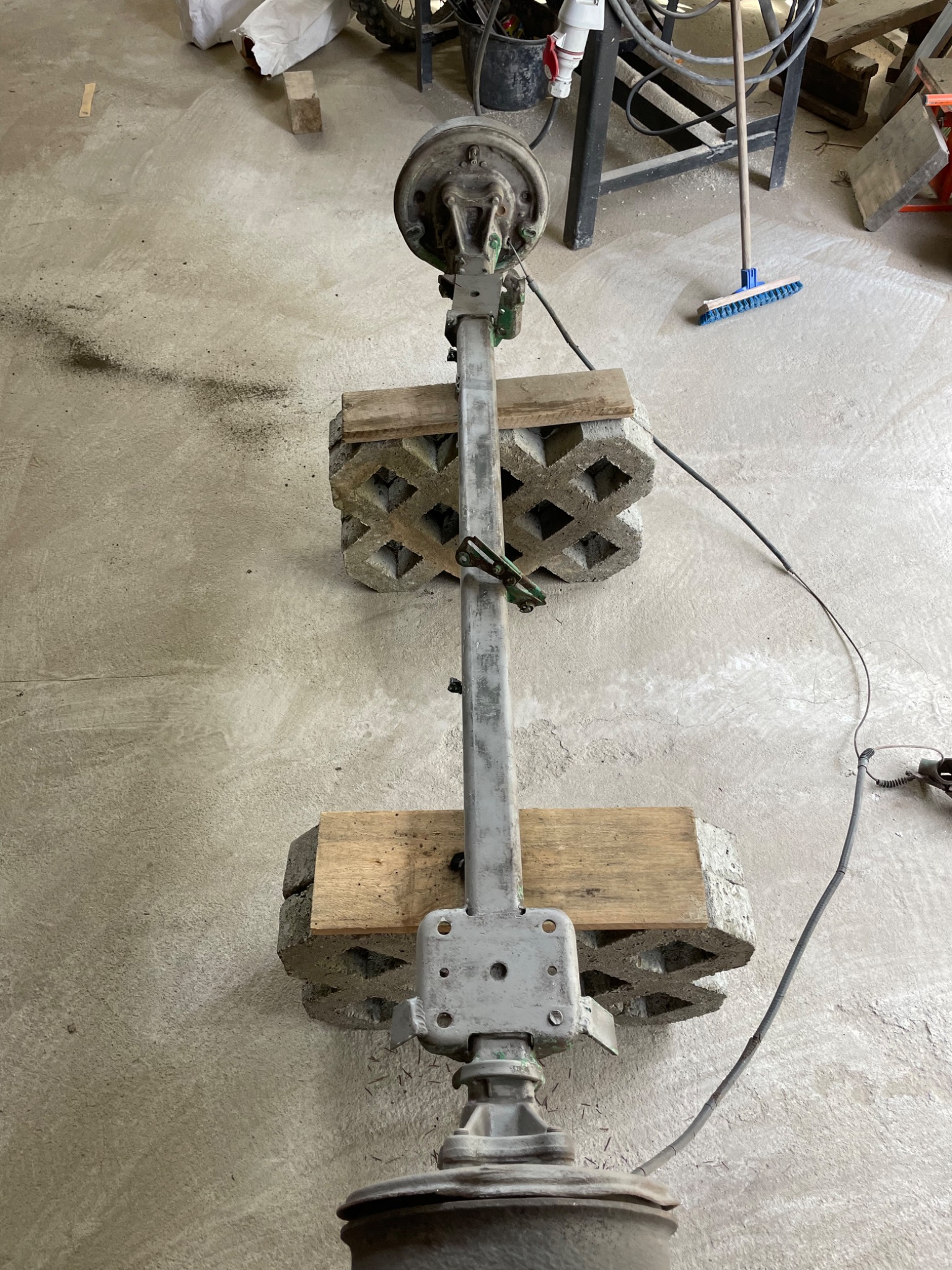 Szara oś z elementami mocującymi, podparta na betonowych pustakach i drewnianych klockach, widoczna w warsztacie na betonowej podłodze.