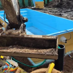 Wykop pod basen z użyciem koparki, widoczny niebieski basen z tworzywa sztucznego, ziemia, rury i liny zabezpieczające.