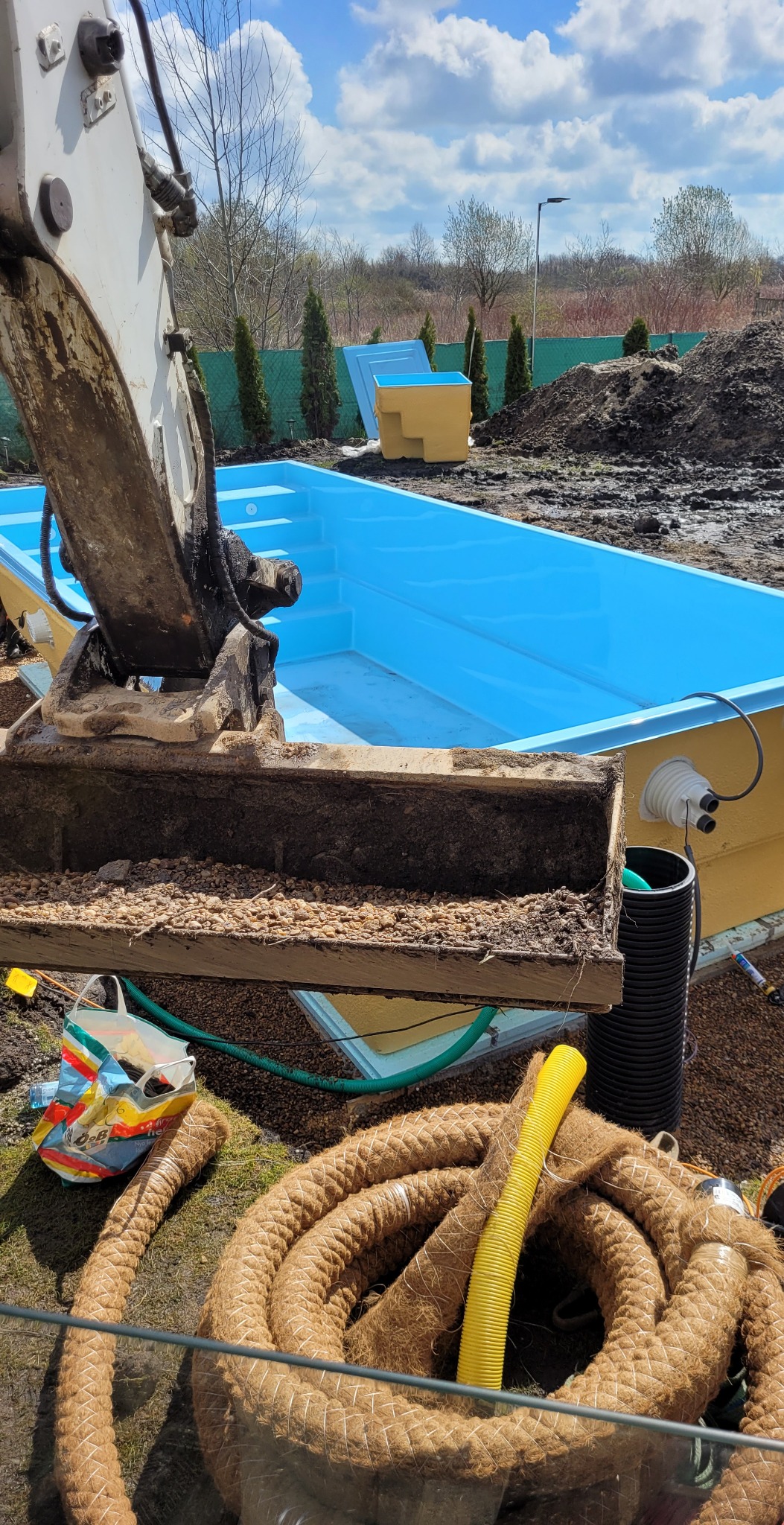 Wykop pod basen z użyciem koparki, widoczny niebieski basen z tworzywa sztucznego, ziemia, rury i liny zabezpieczające.