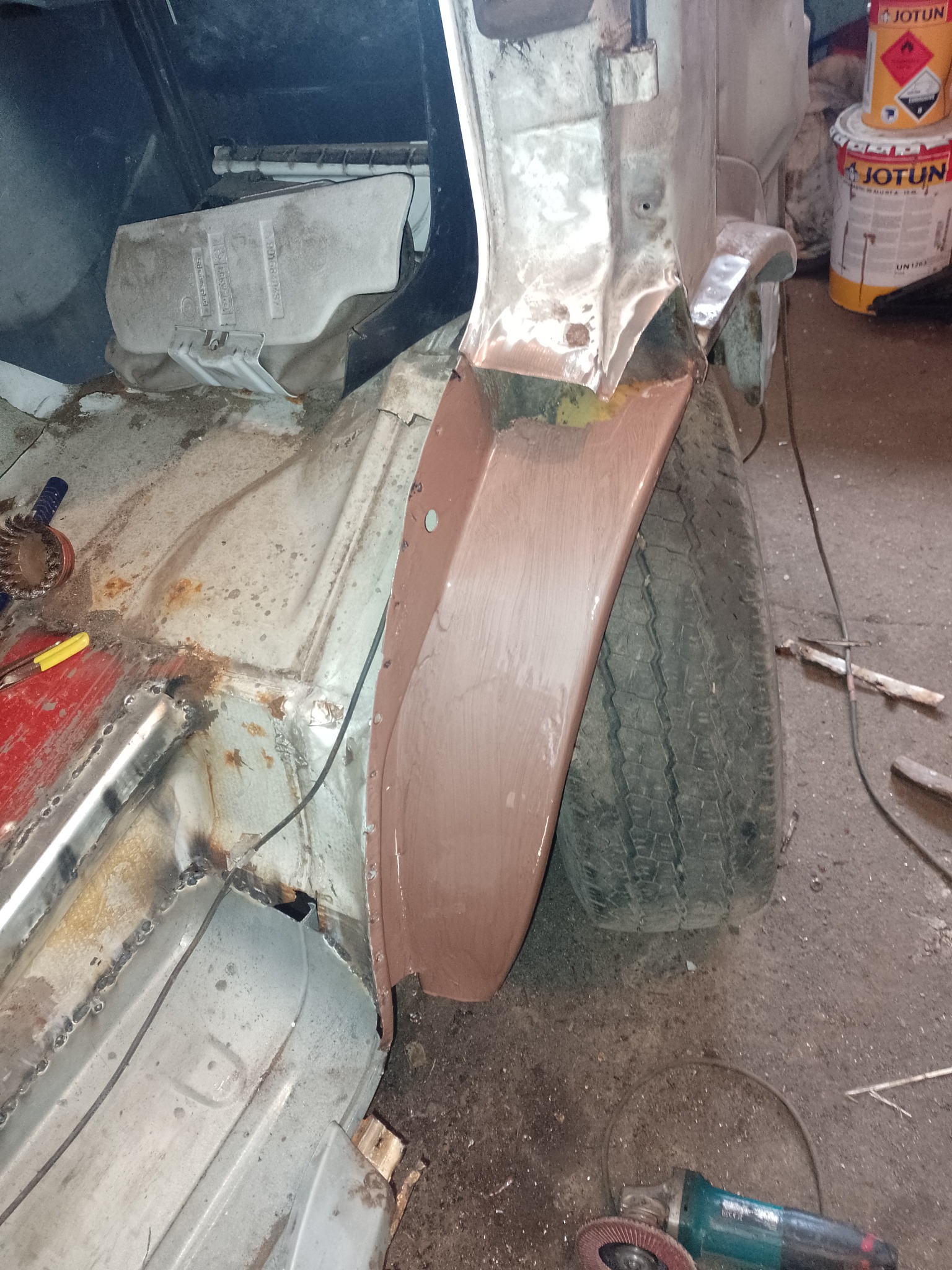 Widok na nadkole samochodu w trakcie naprawy blacharskiej, z widocznymi śladami spawania, szlifowania i nałożoną warstwą podkładu, obok widoczna opona i szlifierka kątowa.