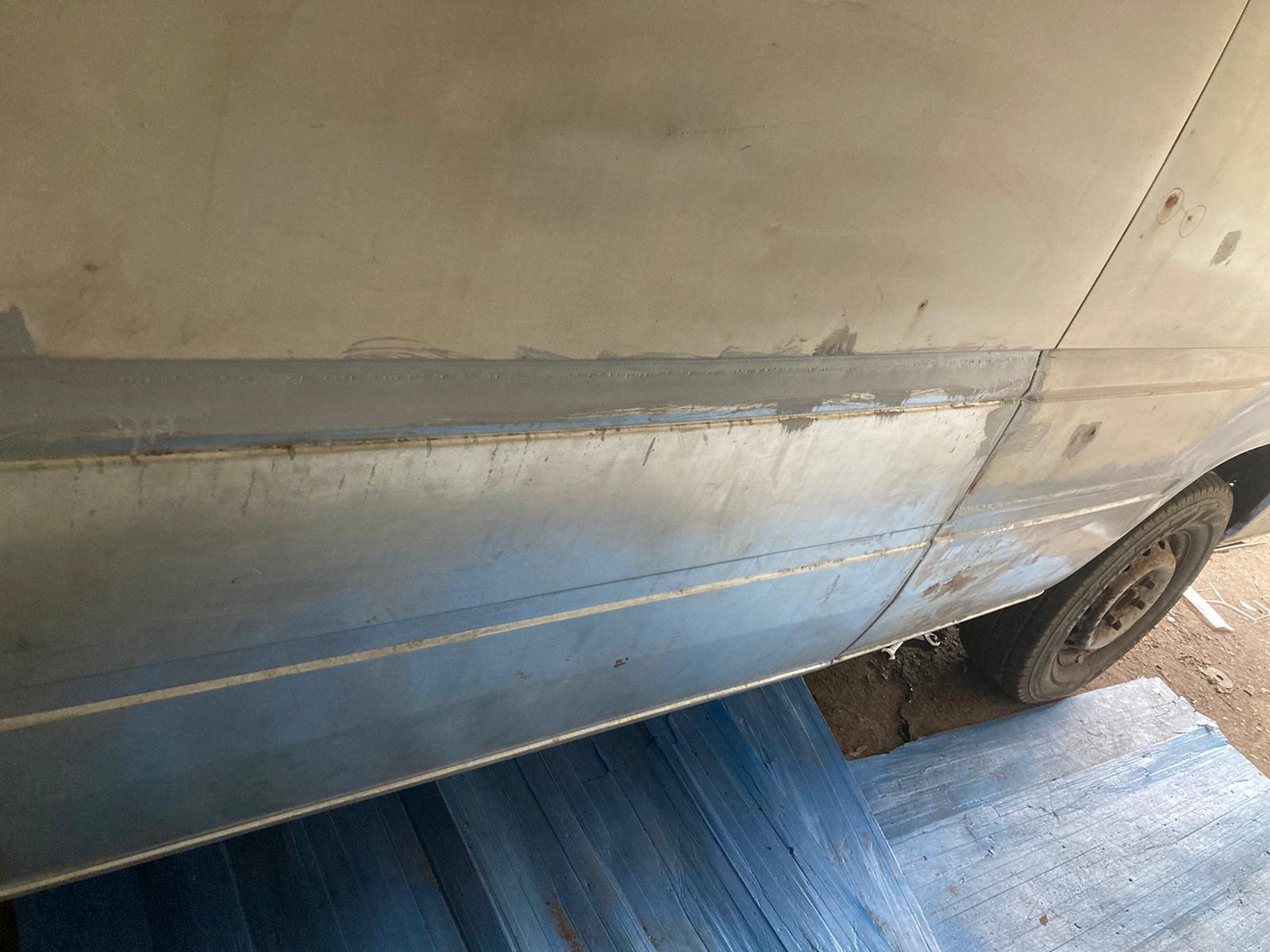 Fragment boku samochodu dostawczego z widocznymi śladami szpachlowania i przygotowania do malowania, na tle niebieskiej, drewnianej platformy.