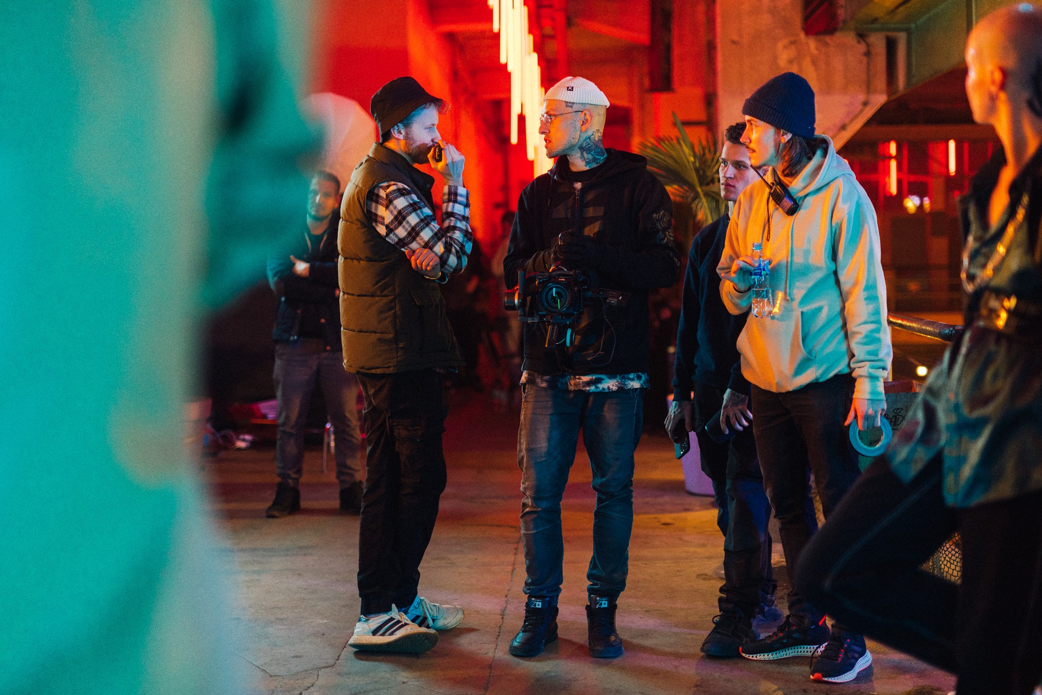 Ekipa filmowa w trakcie pracy na planie, oświetlonym neonowym światłem, kamerzysta z tatuażami na szyi trzyma kamerę, a drugi mężczyzna w czapce i kamizelce gestykuluje podczas rozmowy.