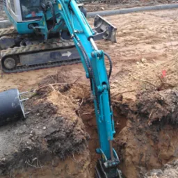 Niebieska mini koparka wykonuje wykop w ziemi, widoczny operator w kabinie, przygotowanie terenu pod instalacje.