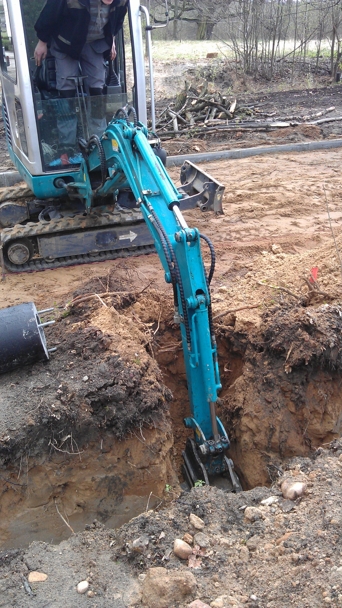 Niebieska mini koparka wykonuje wykop w ziemi, widoczny operator w kabinie, przygotowanie terenu pod instalacje.