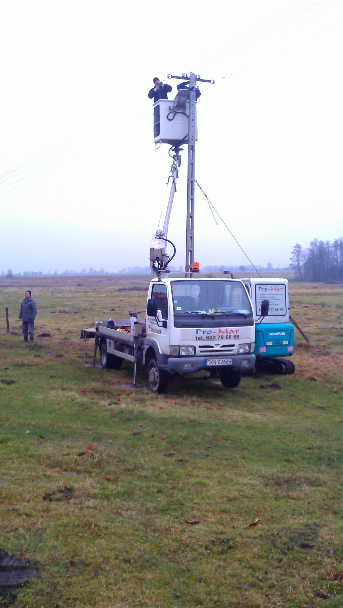 Pracownicy w podnośniku koszowym naprawiający linię energetyczną na słupie, w tle widoczny samochód z podnośnikiem i osoba obserwująca, teren podmokły.