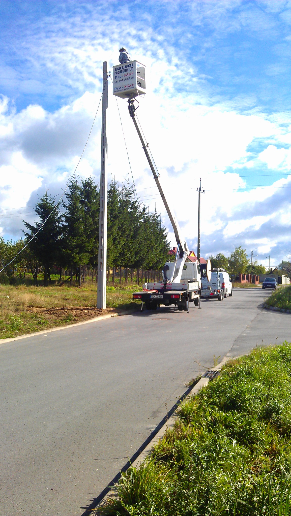 Pracownik w podnośniku koszowym przy słupie energetycznym, widoczny samochód z podnośnikiem i inne pojazdy na drodze, w tle drzewa i niebo z chmurami.
