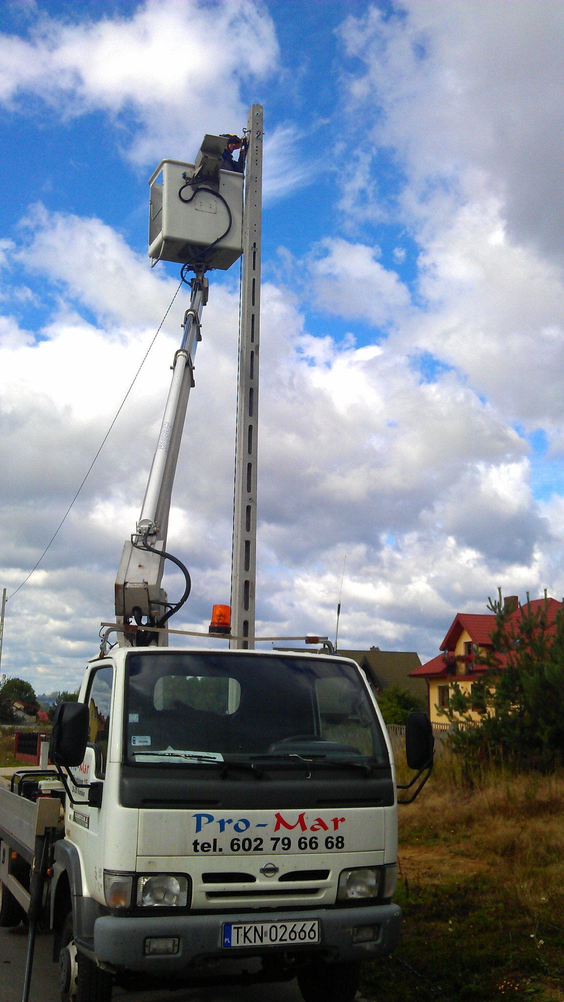 Pracownik na podnośniku koszowym przy słupie oświetleniowym, na tle błękitnego nieba z białymi chmurami, widoczna zabudowa jednorodzinna i firmowy samochód Pro-Mar z numerem telefonu.