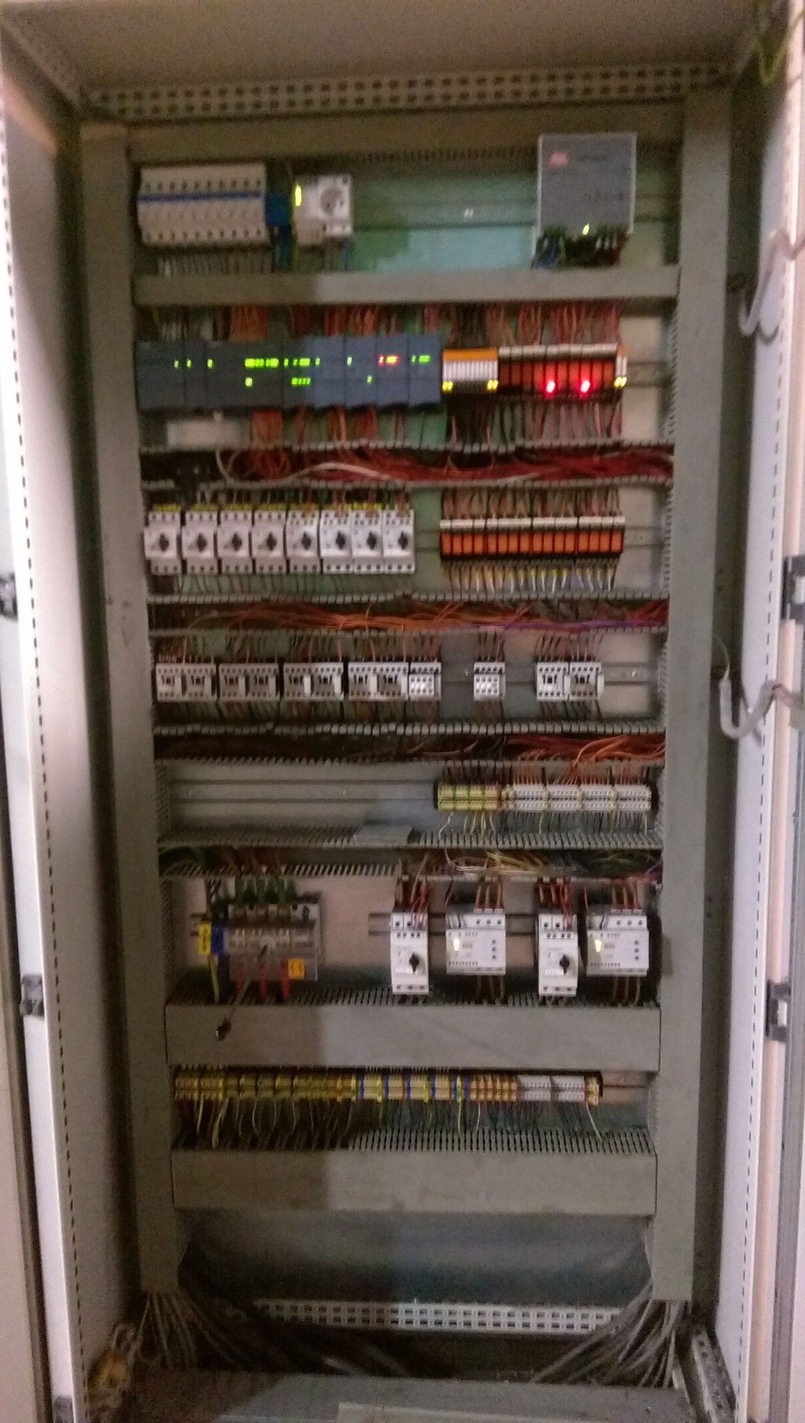 Otwarta szafa sterownicza z widocznymi elementami automatyki, przekaźnikami, bezpiecznikami i okablowaniem, oświetlona od góry, z nieostrymi elementami w tle.