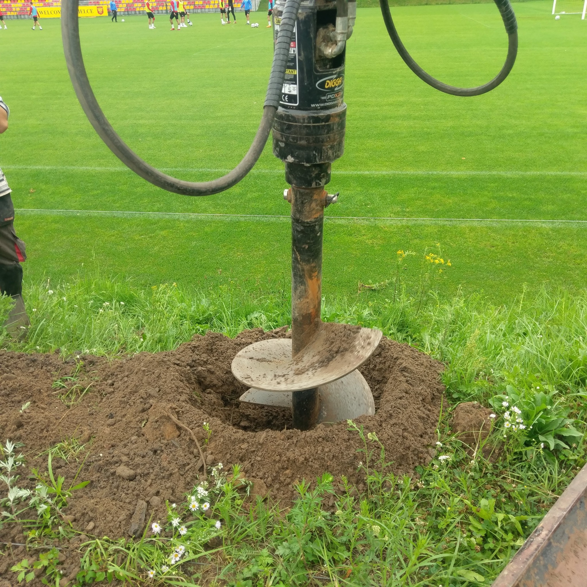 Wiertnica glebowa Digga PDX2 w trakcie wiercenia otworu w ziemi na tle boiska sportowego.
