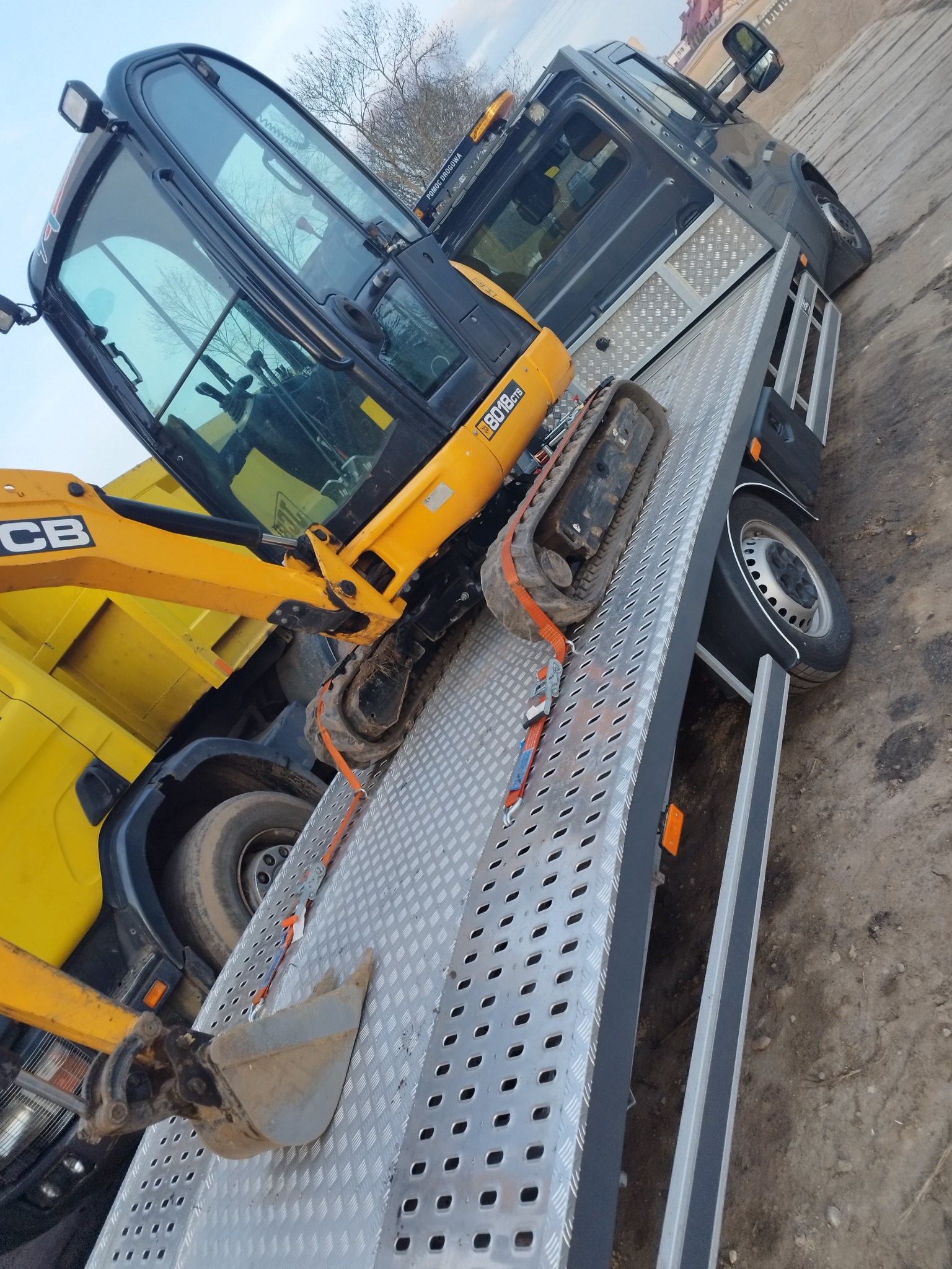 Żółta mini koparka JCB 8018 CTS zabezpieczona pasami na metalowej platformie lawety samochodowej, widok z boku.