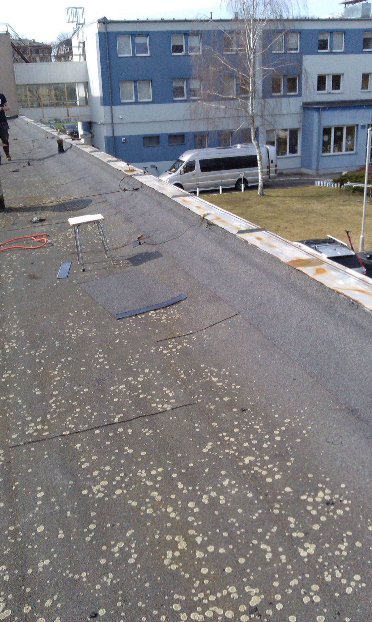 Nisko nachylony dach pokryty papą z widocznymi plamami i fragmentami blachy, w tle budynek z białym busem.