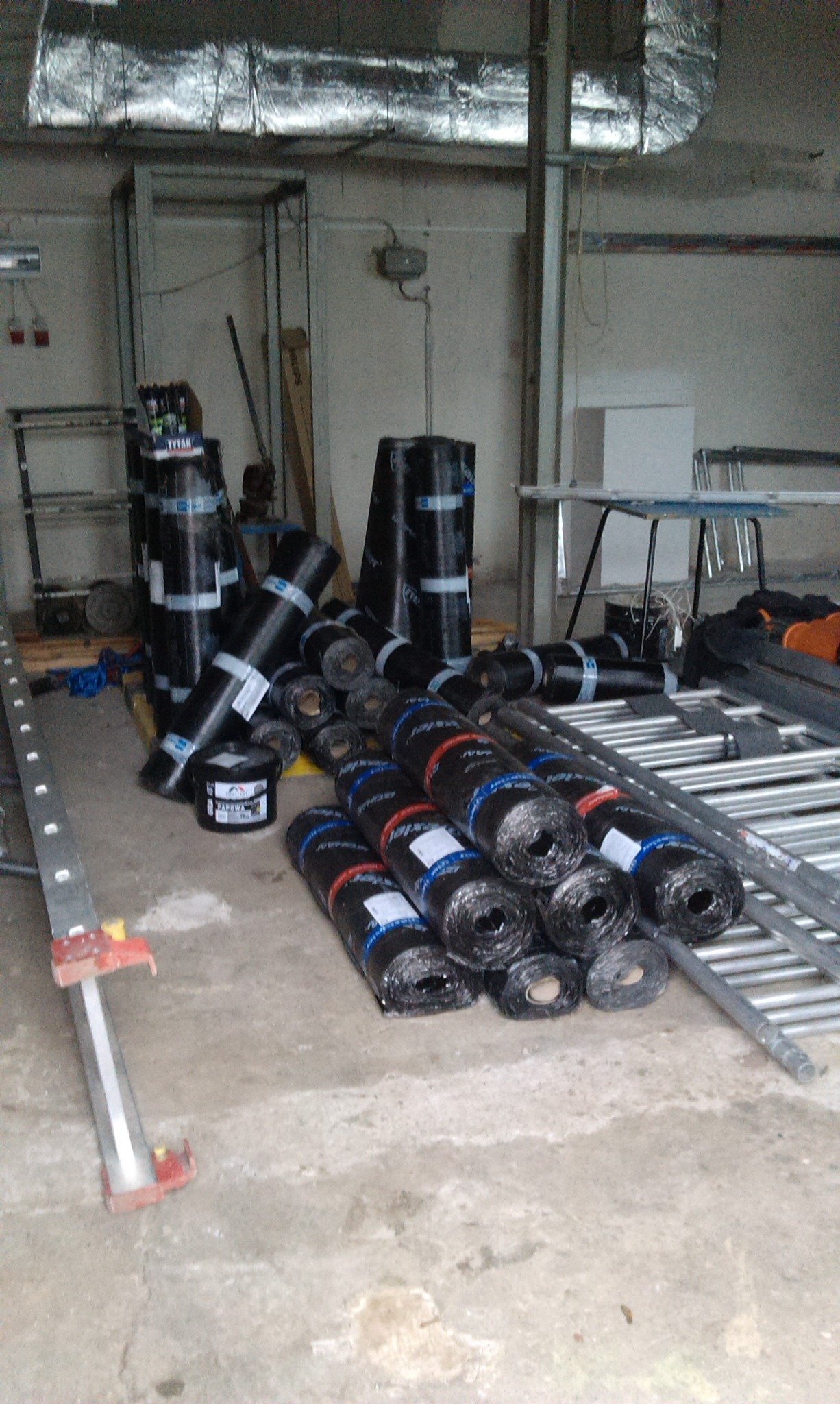 Stos rolek papy termozgrzewalnej w magazynie budowlanym, widoczne etykiety producentów i drabina aluminiowa.