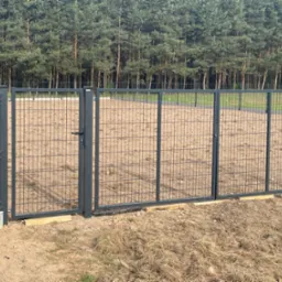 Grafitowa brama panelowa z furtką, zamontowana na drewnianych podkładkach, widok na leśne tło.