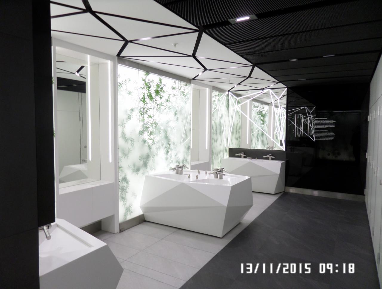Nowoczesna łazienka publiczna z geometrycznymi umywalkami, podświetlanym panelem z motywem roślinnym i czarno-białym sufitem kasetonowym.
